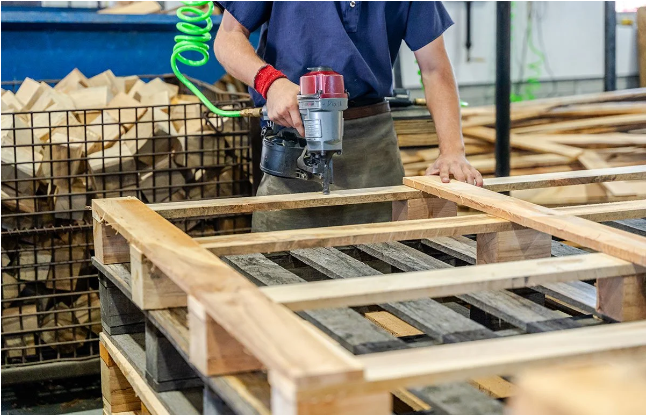 A worker repairing a damaged wooden pallet on a workbench using a power drill