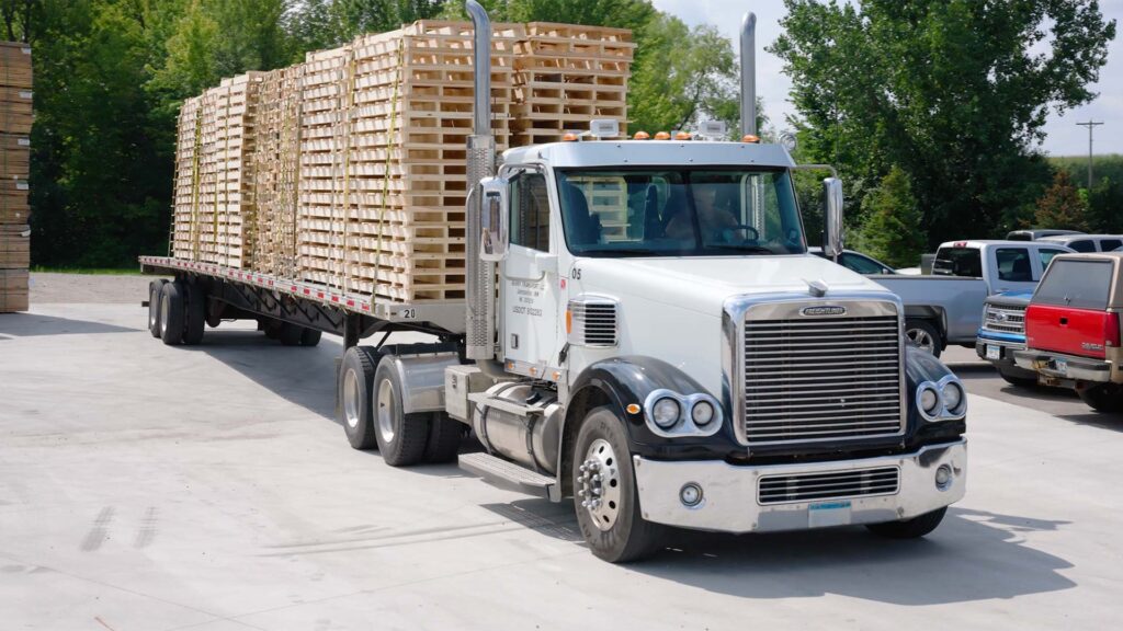 A white Freightliner flatbed semi truck loaded with stacked wooden pallets, parked at the Pallets Pallets Pallets yard ready for delivery